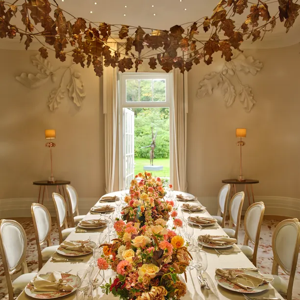 Oak Room set for a social dinner with autumnal floral arrangements and hanging leaf garlands at Coworth Park