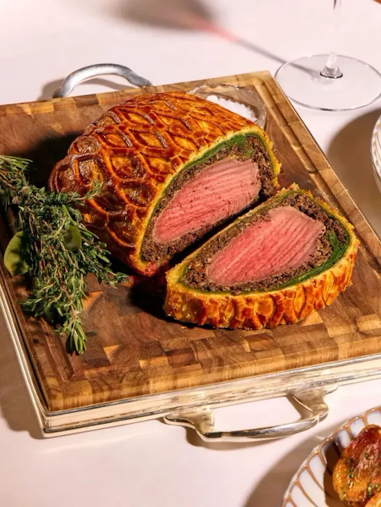 Dining table at The Grill at The Dorchester, showcasing a Beef Wellington sliced to reveal a pink centre, served with asparagus, creamed spinach, roasted potatoes and a fresh seasonal salad.
