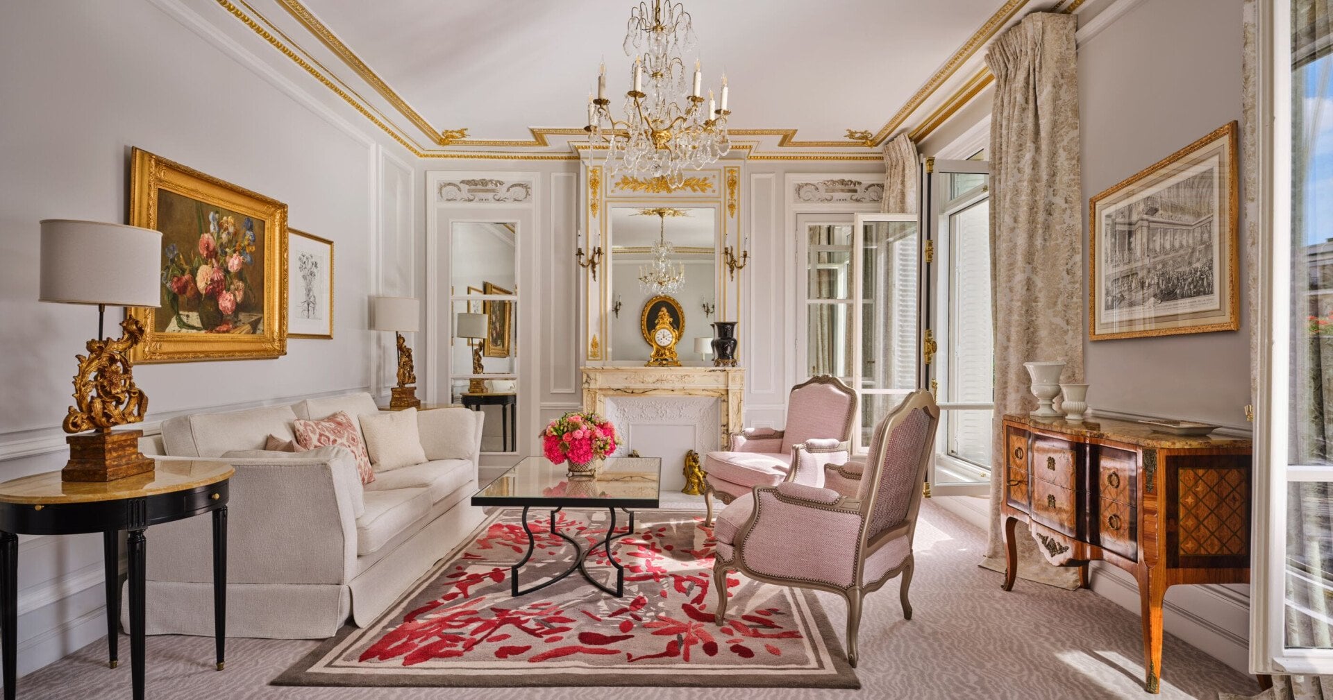Sala de estar elegante com decoração clássica: sofá branco, poltronas cor-de-rosa, lustre ornamentado, detalhes dourados, arte floral e tapete com padrão vermelho. Ambiente aconchegante e luxuoso.