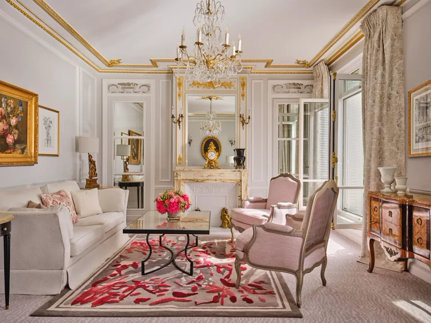 Salon élégant à la décoration classique : canapé blanc, fauteuils roses, lustre orné, touches dorées, compositions florales et tapis à motifs rouges. Ambiance chaleureuse et luxueuse.