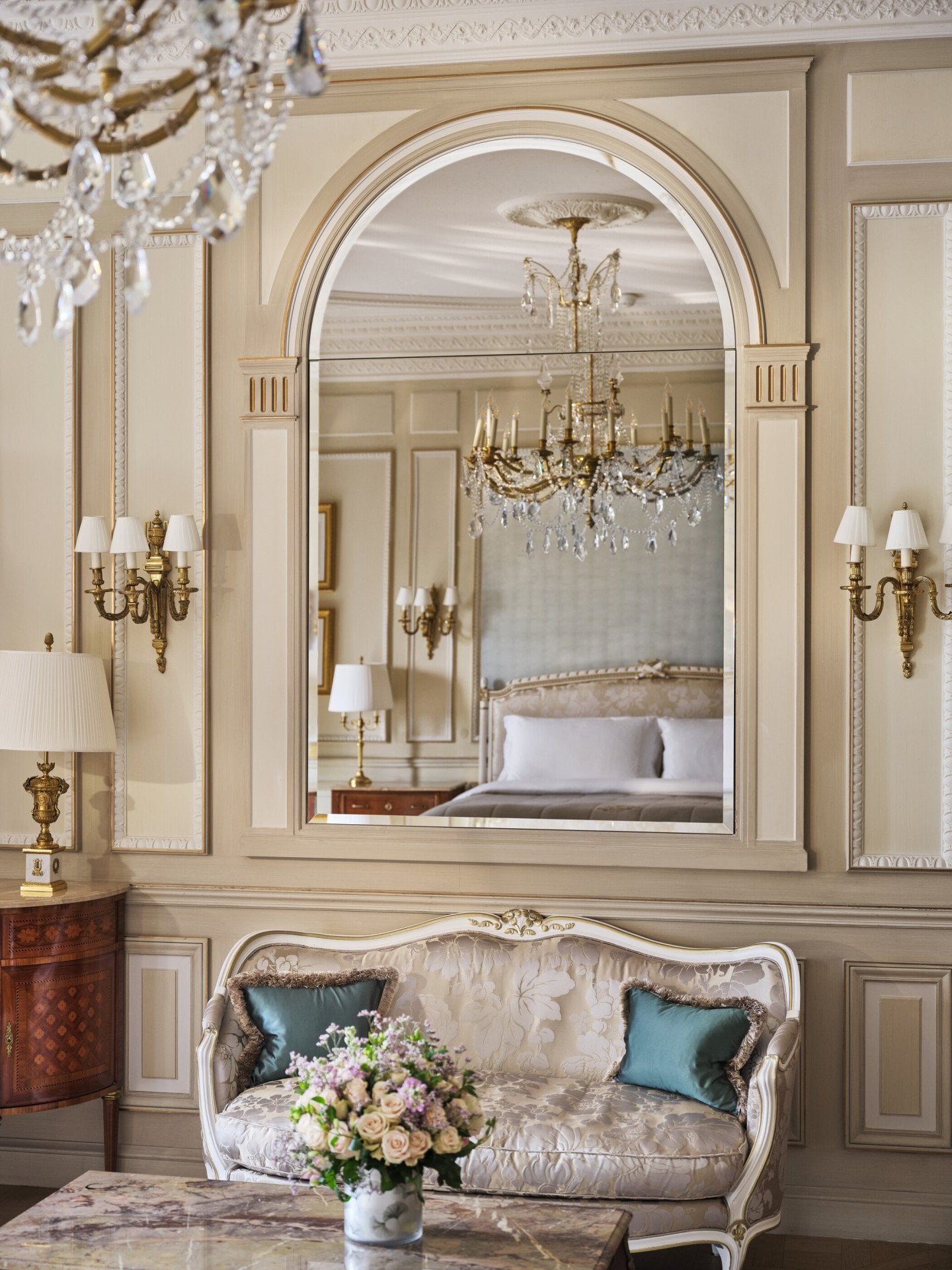 Vista sobre a mesa feita com marchetaria de pedra e um sofá com espelho acima, que reflete o lustre e a cama, no Le Meurice, Paris