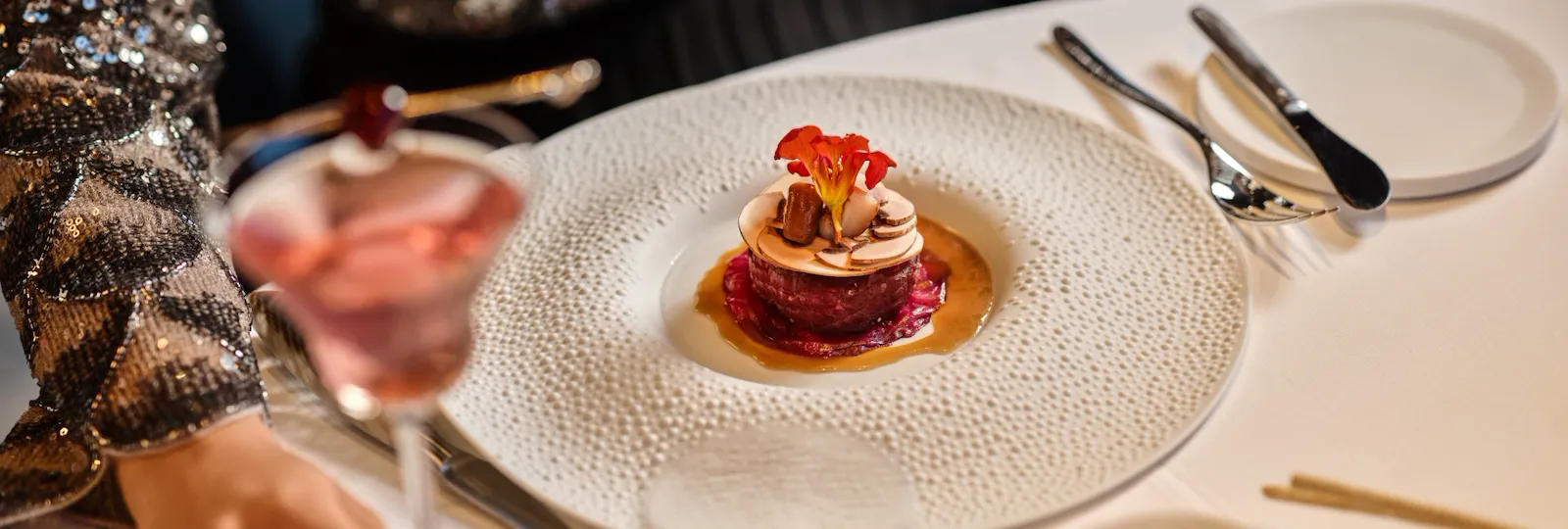 Gourmet dish of beef placed on an elegant table settign in front of a woman holding a drink