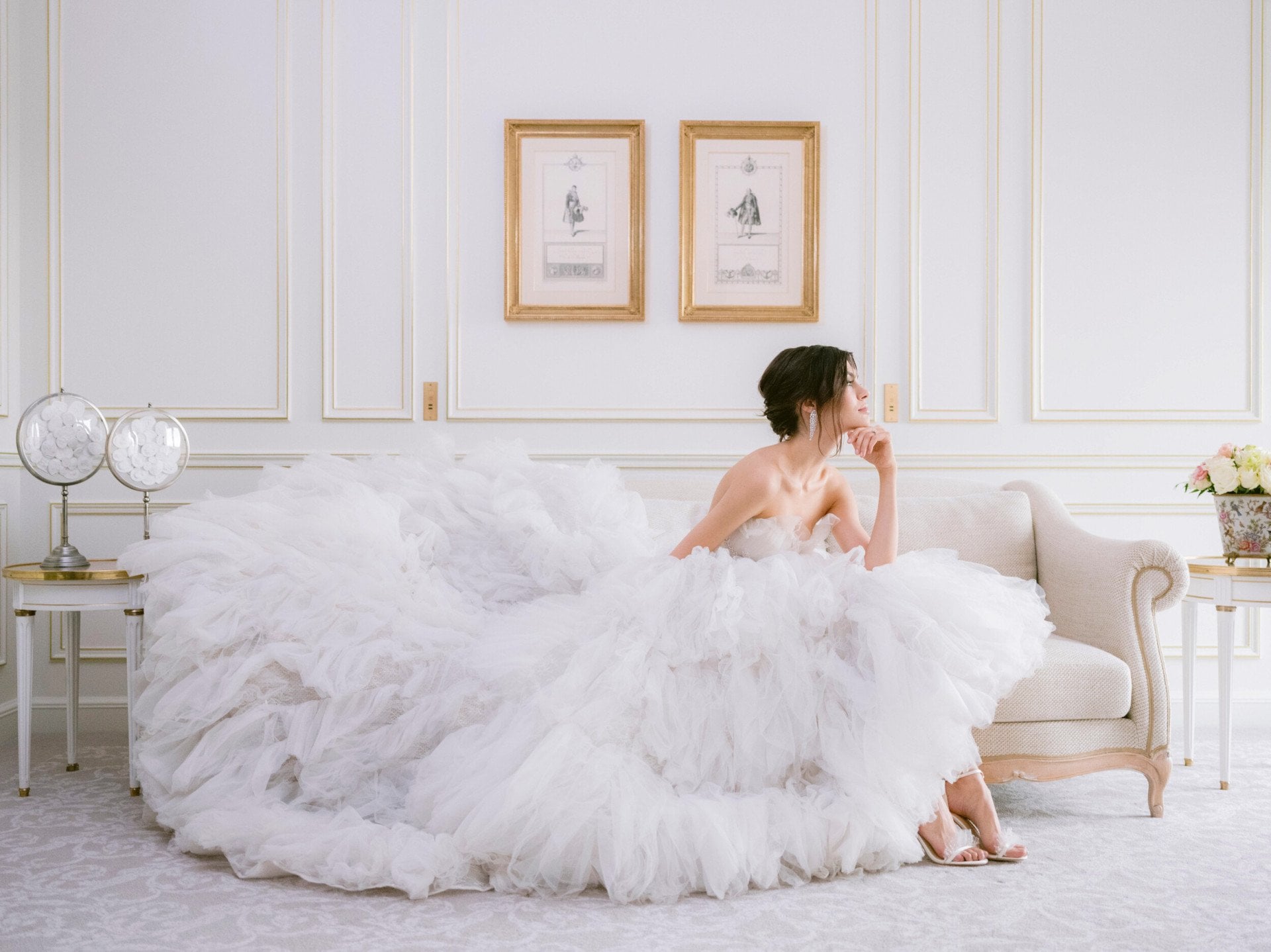 Noiva com um grande vestido branco com tecidos delicados, sentada numa suite num sofá e cobrindo-o com o seu vestido, enquanto olha para fora da janela com um olhar sonhador. Na parede, há dois desenhos em molduras douradas, o sofá é bege claro e o chão está coberto com um tapete estampado cinzento/prateado, no Le Meurice, Paris.