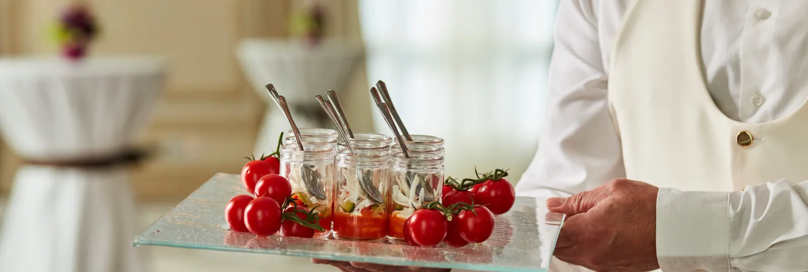 A close-up of a tray of canapés and decorative tomatoes being carried by a waiter. In the background, two high tables.