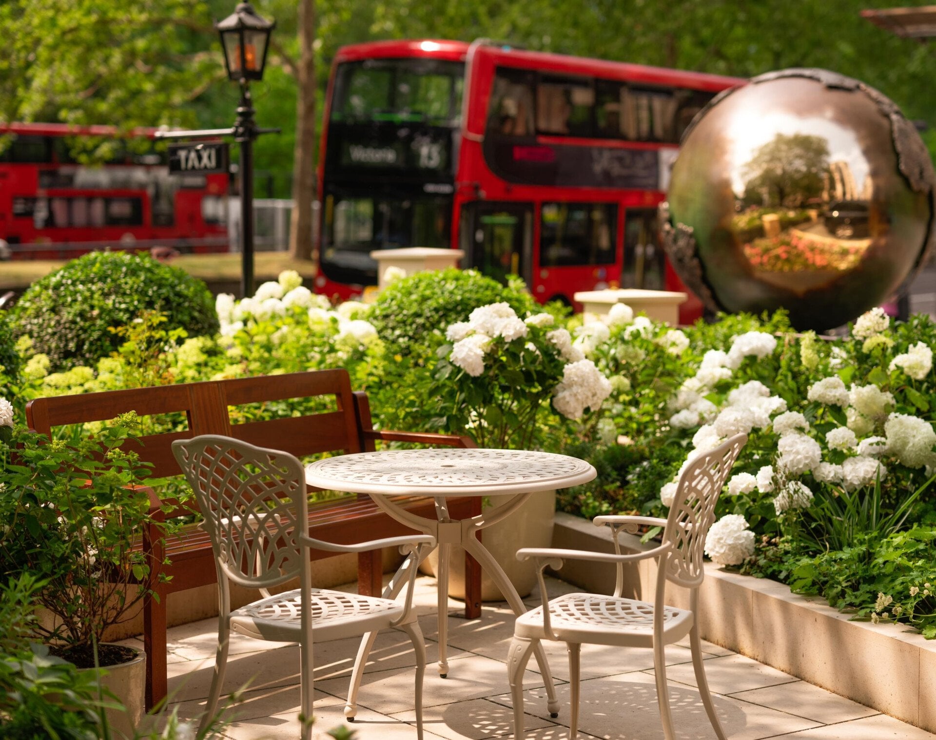 The Dorchester’s outdoor garden showcasing the globe sculpture, elegant al fresco seating with views of Hyde Park and a red London bus travelling along Park Lane.
