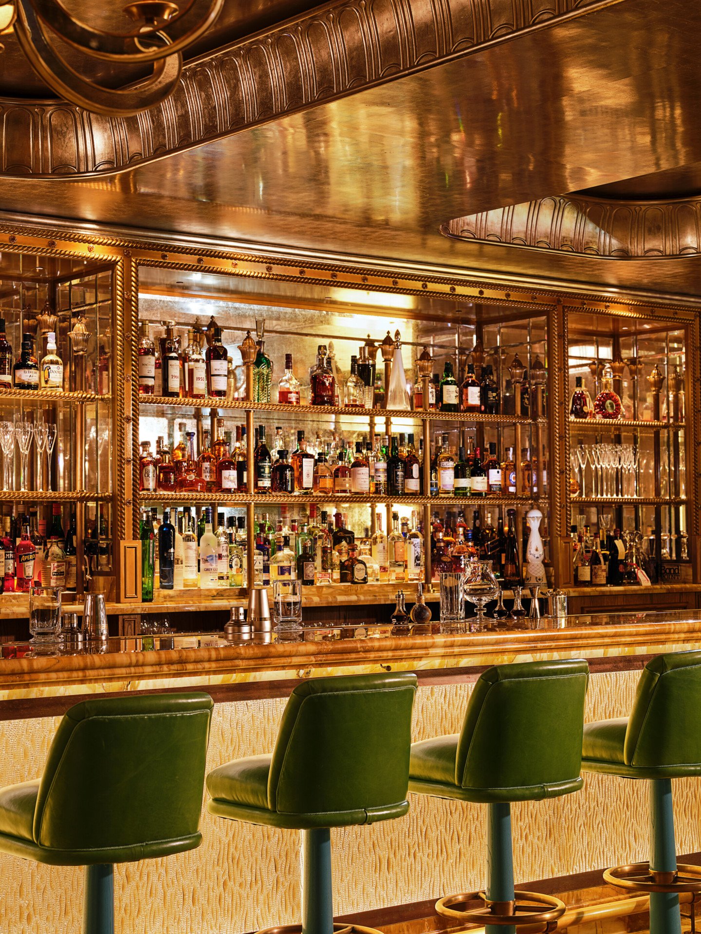 Green leather bar stools line the marble bar at The Vesper Bar, with illuminated shelves displaying an extensive collection of spirits.