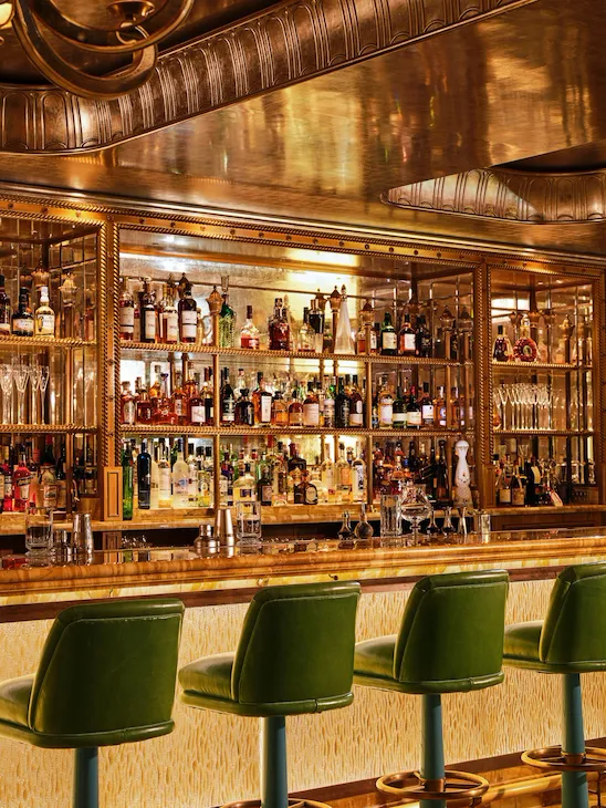 Green leather bar stools line the marble bar at The Vesper Bar, with illuminated shelves displaying an extensive collection of spirits.