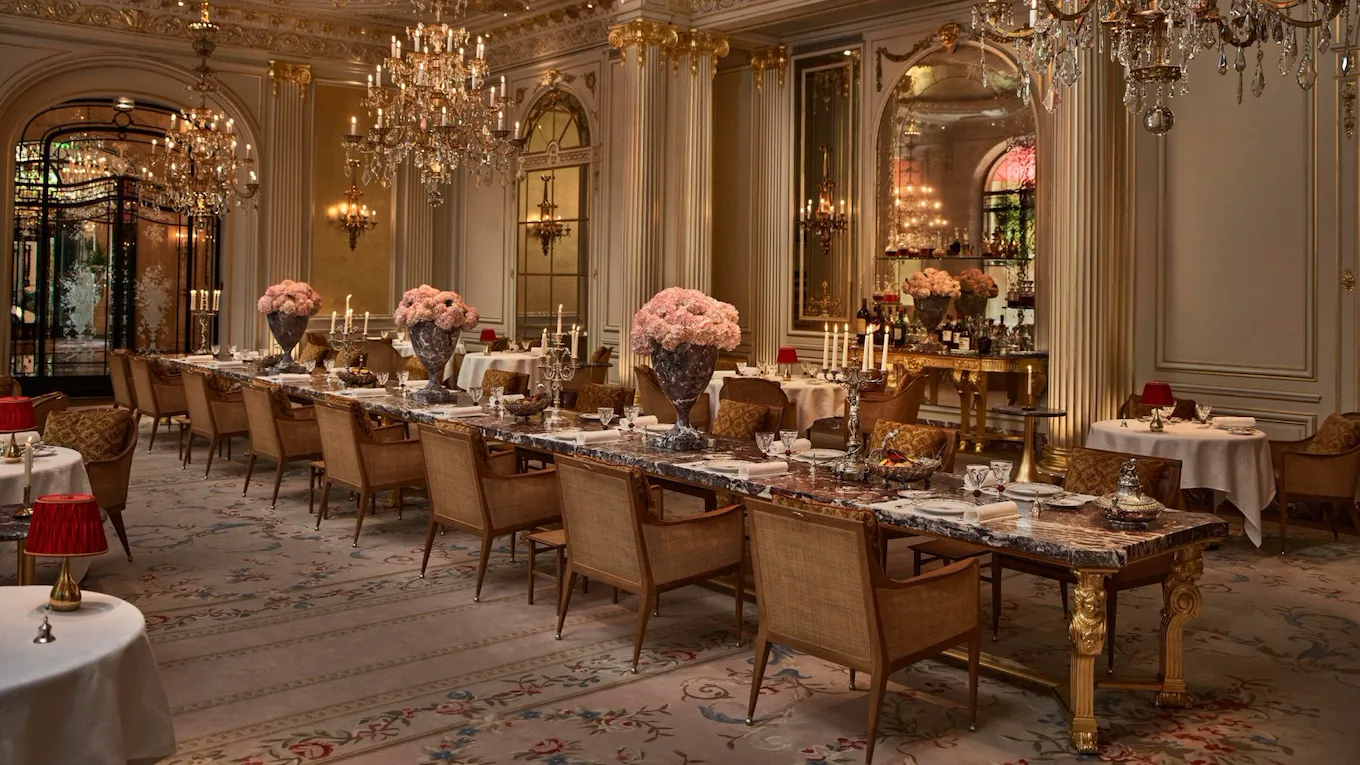 Overview of the Jean Imbert au Plaza Athénée Dining Room and its central marble table with flowers and crystal chandeliers hanging from the ceiling. At Hotel Plaza Athénée, Paris