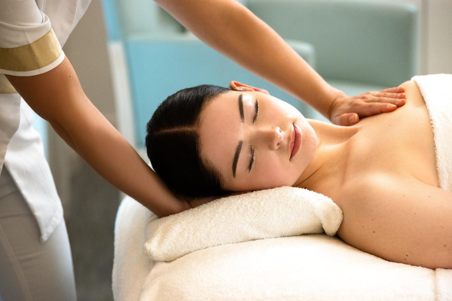 We can see a woman lying on a massage table, resting her head on a white pillow. We can see the client's face positioned on her side. She has her eyes closed and her hair is tied back. On the left-hand side of the photo, we can see the masseur's uniform. One of the hands is positioned under the client's head and the second hand is on her upper chest. We can see the white blanket covering the client's chest at Le Meurice, Paris.
