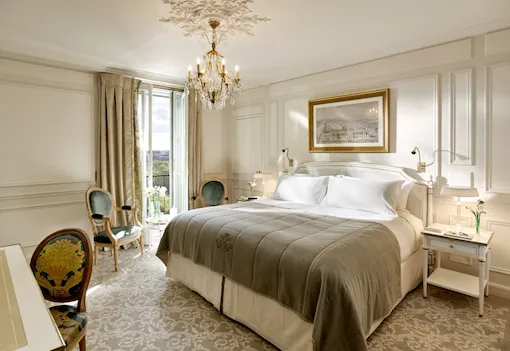 Global view of an executive room highlighting the light grey and cream undertones, as well as the view over the Tuileries gardens in the background. Jewelry and books are on the bedside tablesAt Le Meurice, Paris.
