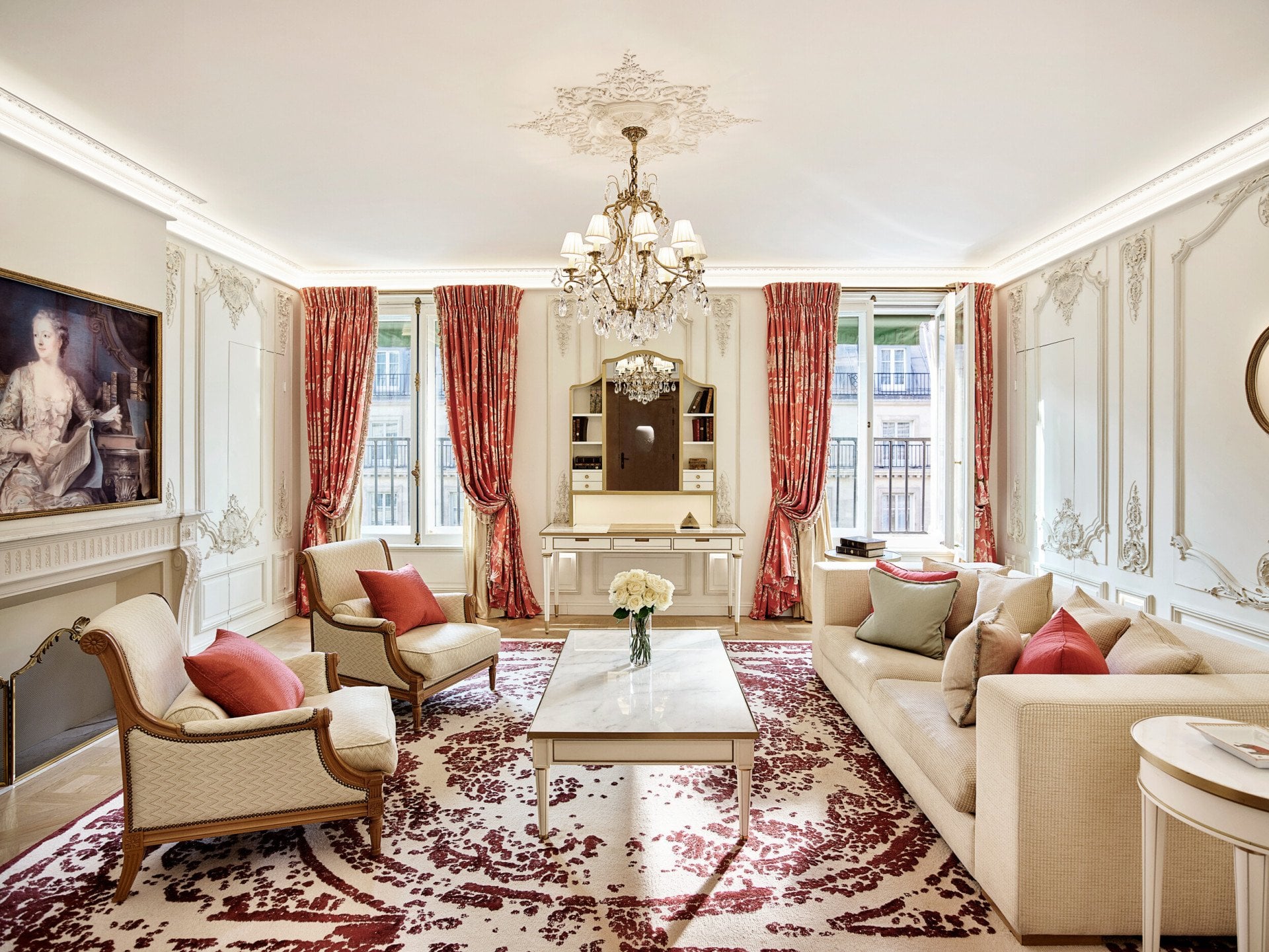 Sala de estar luxuosa com lustre ornamentado, sofás elegantes e detalhes em vermelho. Janelas amplas com cortinas vermelhas, tapete floral e pintura clássica.