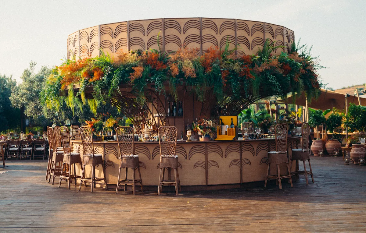 A round outdoor bar features tall wicker chairs and lush green and orange foliage. It stands on a wooden deck amid an inviting, sunlit garden setting.