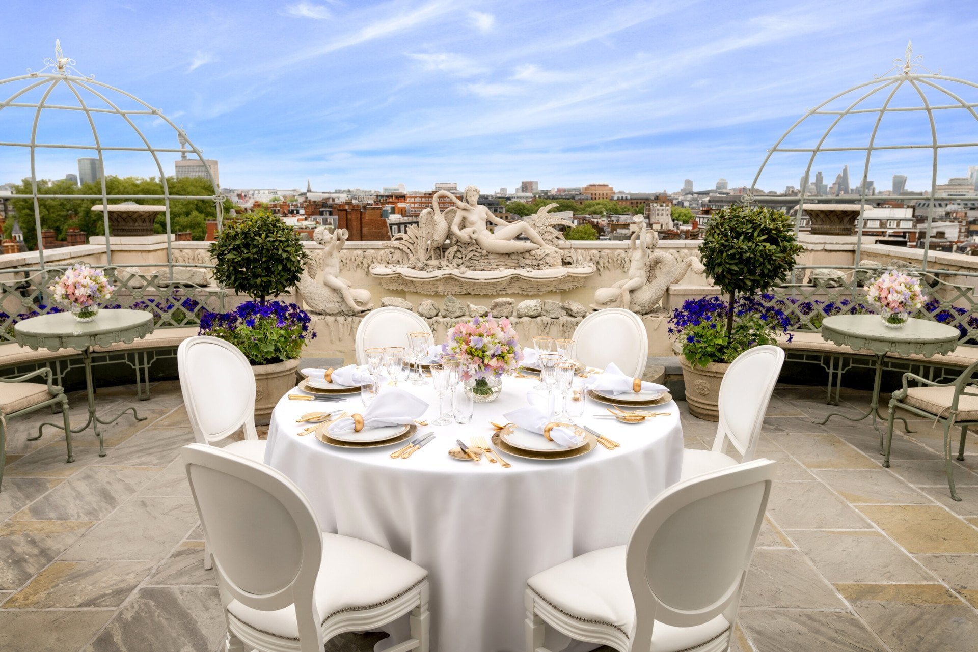 Luxurious rooftop terrace at The Penthouse & Pavilion at The Dorchester, showcasing a round table elegantly laid for dining, surrounded by white chairs, seasonal flowers, classical stone statuary, manicured topiary and panoramic views of the London skyline beneath a blue sky.