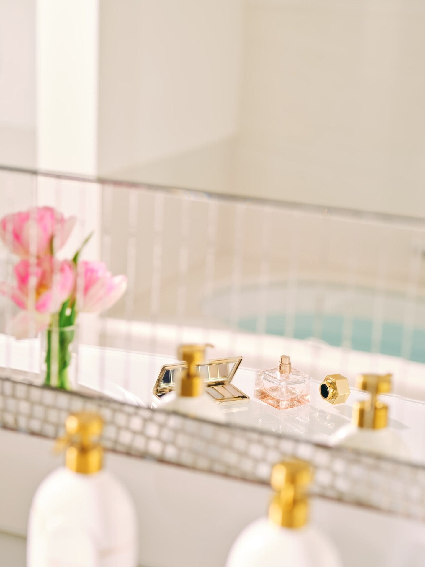 riflesso dello specchio di un bagno con fiori rosa, un profumo, una palette di trucchi e dei dispenser di shampoo
