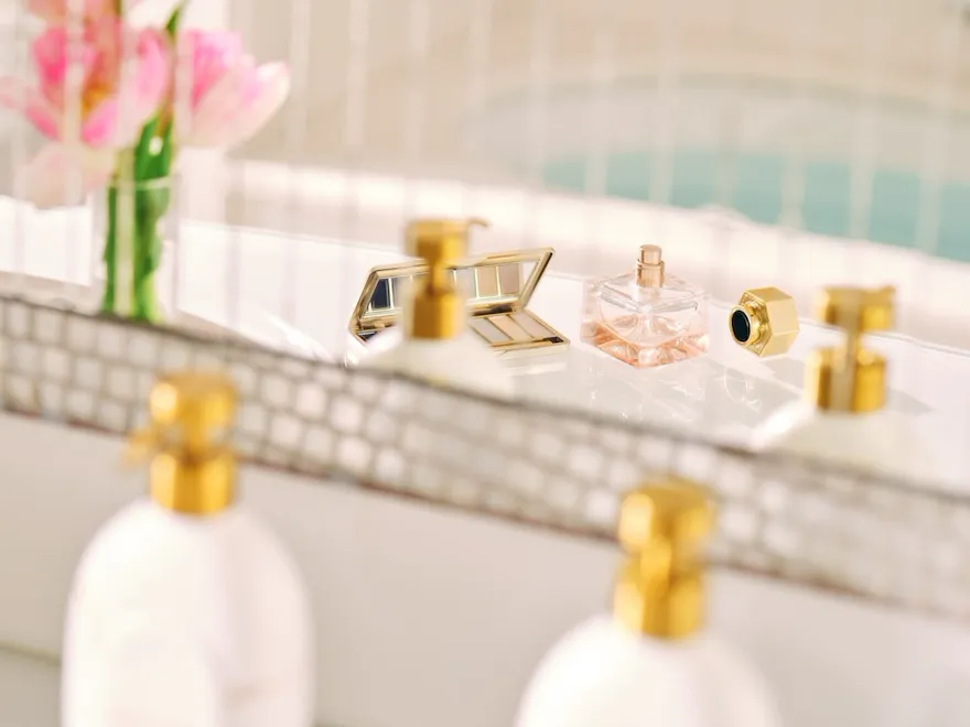 riflesso dello specchio di un bagno con fiori rosa, un profumo, una palette di trucchi e dei dispenser di shampoo