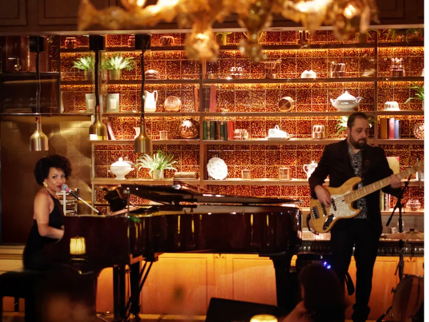 Ronnie Scott’s at The Dorchester, showcasing a live performance with musicians playing piano and bass, framed by warm lighting and backlit shelves displaying decorative objects and glassware.