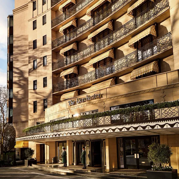 Exterior view of The Dorchester, showcasing its elegant Park Lane façade, decorative wrought-iron balconies, signature yellow-striped awnings and the hotel's welcoming entrance bathed in warm sunlight.
