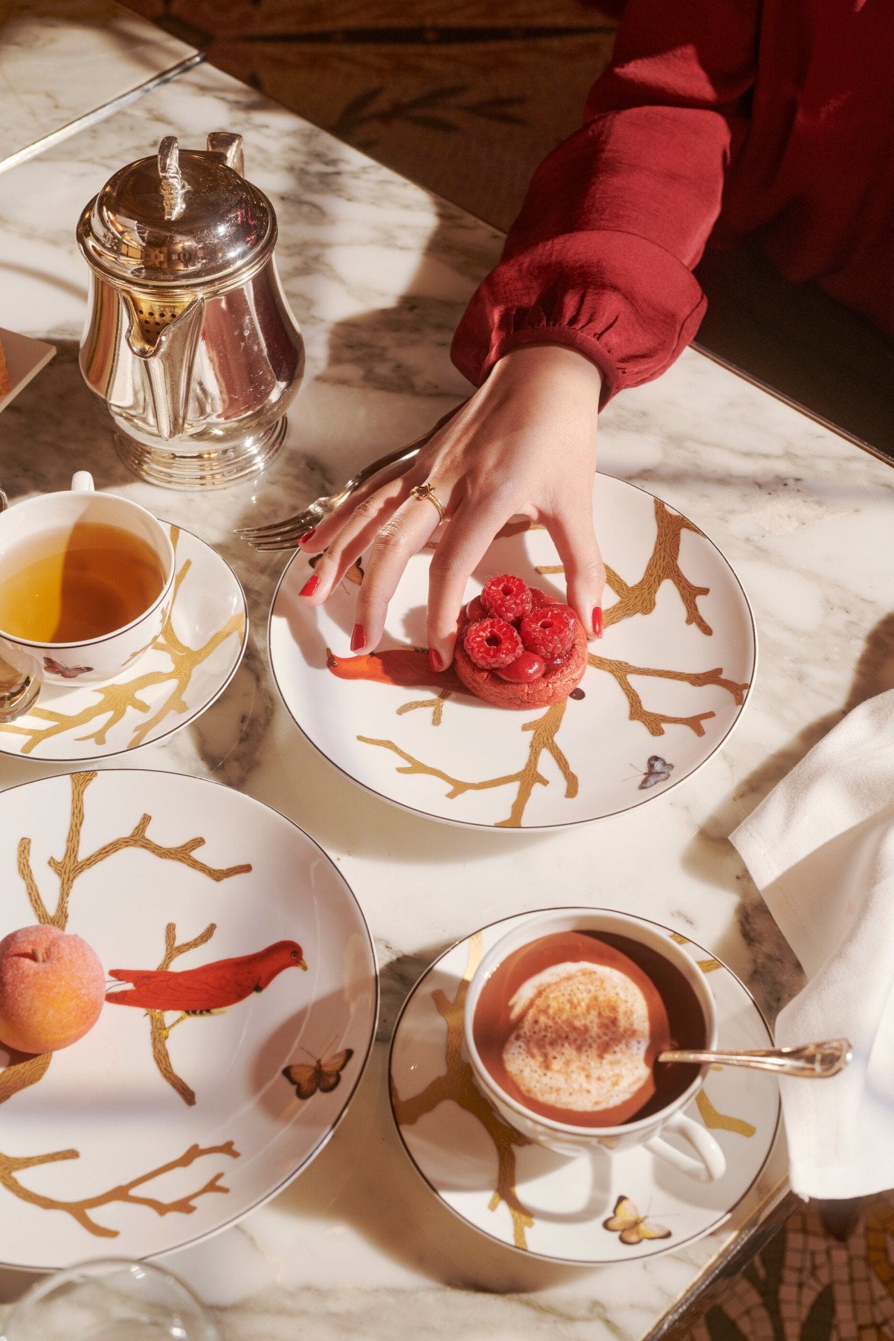 Alguns pormenores da hora do chá com uma mão a tirar uma bolacha de framboesa de Cédric Grolet ao lado de bebidas quentes e um pêssego, no Le Meurice, Paris  