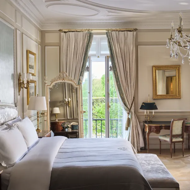 Vista ampla do lado direito da cama com o espelho, a escrivaninha, a cadeira e as janelas abertas atrás, no Le Meurice, Paris
